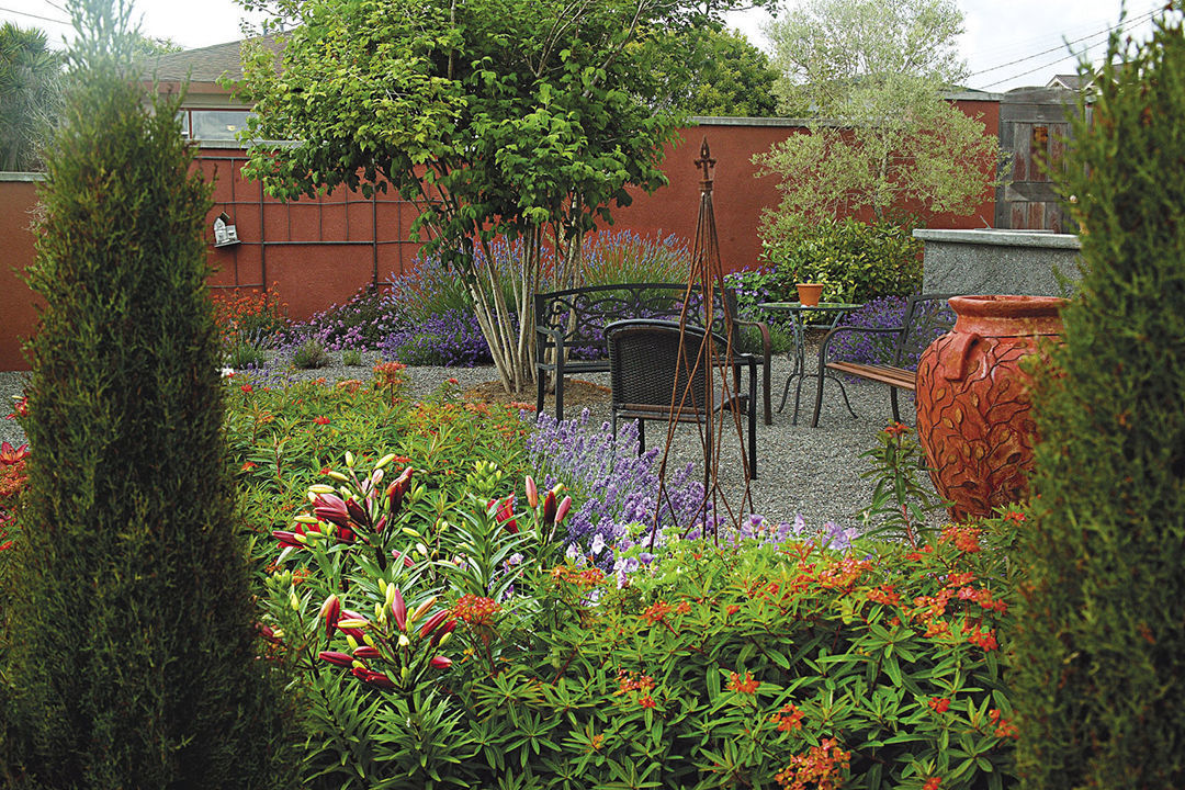 A pair of Italian cypresses in the garden, with benches, a small table, pots and other colorful flowers nearby